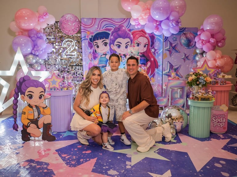 Mujer de vestido claro sonriente junto a niño con camiseta blanca y niña en abrigo colorido, decoraciones festivas de fondo.