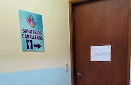 Puerta de baño de madera con letrero de 'Clausurado' en un pasillo de hospital de colores claros y azul.