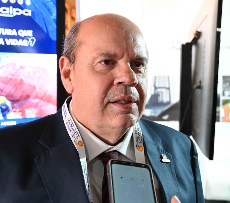 Hombre en traje oscuro y corbata de rayas hablando por teléfono en un evento, con pantalla de fondo que reza 'malpa'.