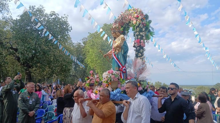 Hombres llevando imagen de la Virgen decorada con flores, rodeados de asistentes en ambiente festivo con banderines azules y blancos.