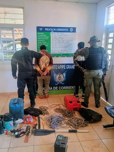 Tres personas en detención en oficina policial, dos con uniformes tácticos oscuros y uno en camiseta clara, rodeados de objetos sospechosos.