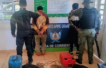 Tres personas en detención en oficina policial, dos con uniformes tácticos oscuros y uno en camiseta clara, rodeados de objetos sospechosos.