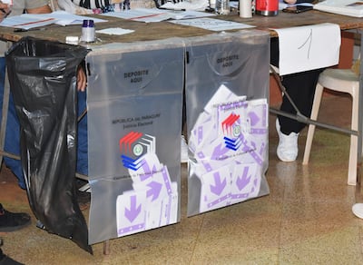 Mesa de votación con urnas transparentes y documentos electorales, en ambiente interior de votación.