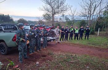 Cuatro agentes armados en uniforme camuflado, rodeados de otros policías, en un entorno rural con árboles y vehículos.