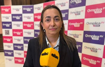 Mujer con cabello largo frente a un micrófono con logo de ABC Color, vestida con traje oscuro y blusa a rayas, en un fondo de panel colorido.