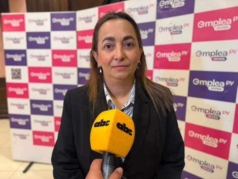 Mujer con cabello largo frente a un micrófono con logo de ABC Color, vestida con traje oscuro y blusa a rayas, en un fondo de panel colorido.