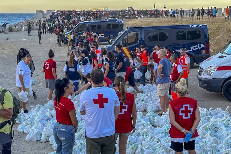 Miembros de la Cruz Roja de Ceuta reparten bolsas de comida a personas migrantes este martes, en la playa El Trampolín de Ceuta. Más de medio millar de migrantes, sobre todo subsaharianos, permanecen en el exterior del Centro de Estancia Temporal de Inmigrantes (CETI) de Ceuta para poder ocupar una plaza en este recinto, que está colapsado al haber cubierto sus 512 plazas. Los migrantes han sido desplazados a la playa del Trampolín, situada a apenas quinientos metros del CETI, con la intención de que no se produzcan incidentes entre estas personas y las que están en el interior del recinto. 