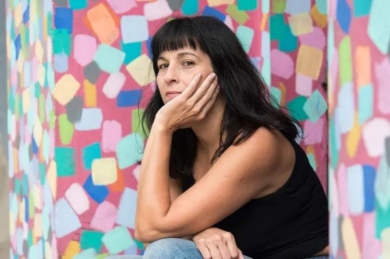 Mujer con cabello largo y oscuro en camiseta negra, sentada en espacio iluminado con fondo colorido y artístico.
