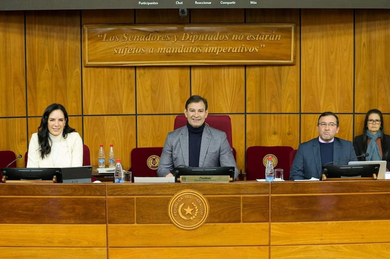 Claudia Centurión vestida de blanco sonríe mientras discute con otros legisladores en ambiente formal con decoraciones en madera.