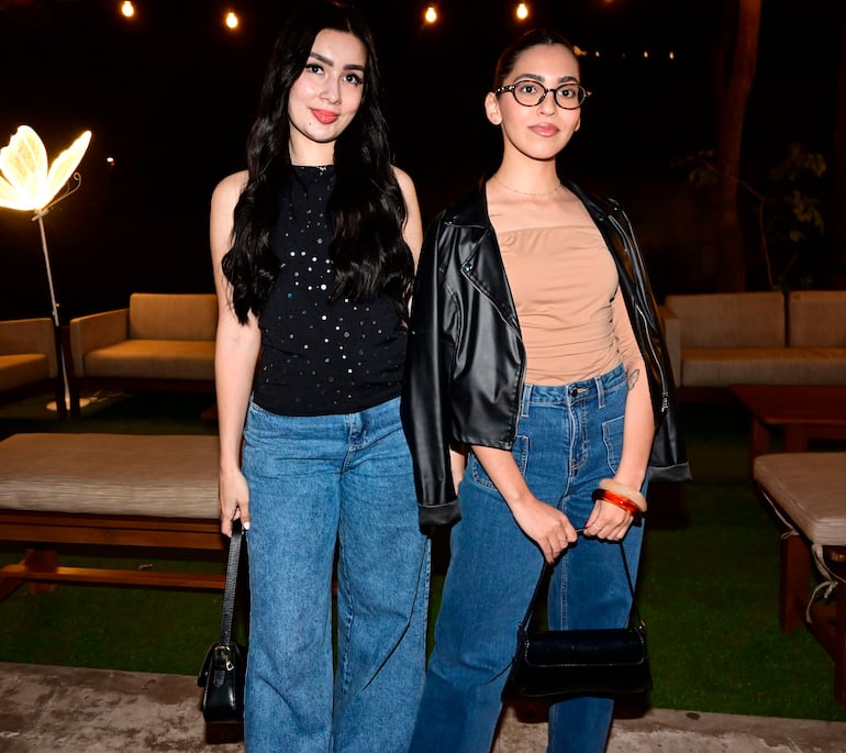 Dos mujeres sonrientes, una con blusa negra y otra con chaqueta de cuero, posan en un evento social con sofás de fondo.