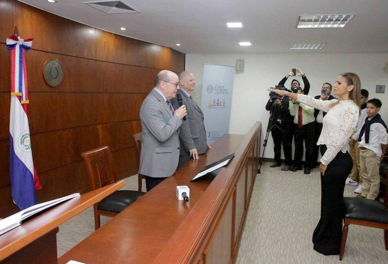 César Diesel habla con micrófono, mientras Luis María Benítez Riera escucha y Natalia Cacavelos levanta la mano en juramento.