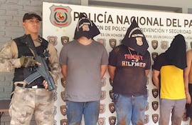 Un agente de policía con arma presenta a cuatro hombres encapuchados, algunos muestran ropa sucia y manchas visibles.