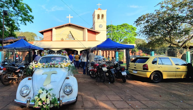 Automóvil blanco decorado con flores frente a la iglesia, con asistentes agrupados esperando para entrar al evento.