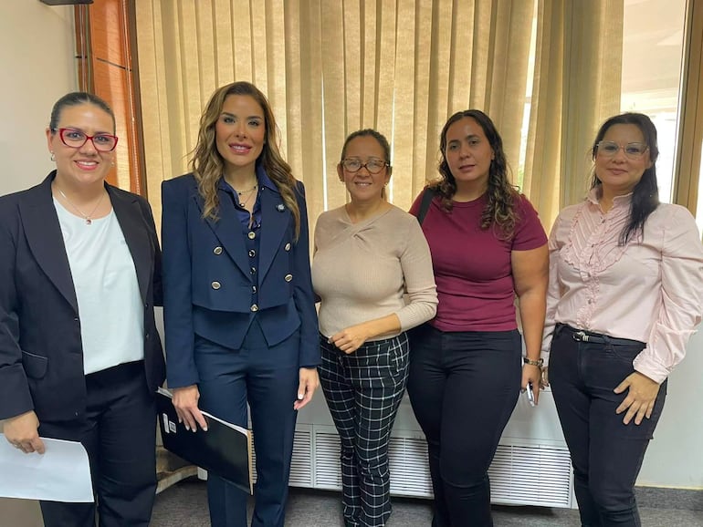 Cinco mujeres vestidas formalmente, de pie, sonrientes en un ambiente profesional con luz natural. La mujer del centro lleva chaqueta negra.