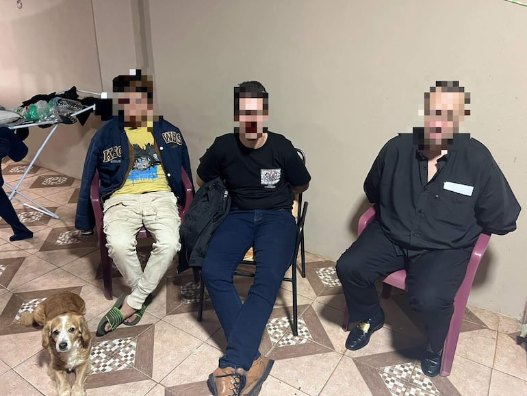 Tres hombres sentados en sillas, uno de ellos al centro. Un perro pequeño está en el suelo frente a ellos en un entorno doméstico.
