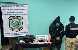 Hombre con abrigo negro y capucha en posición de sumisión, acompañado por un oficial de policía en uniforme antimotines, rodeado de evidencia en una oficina.