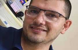 Hombre con gafas y barba ligera, sonriendo en una oficina, con camiseta oscura y fondo de objetos diversos.