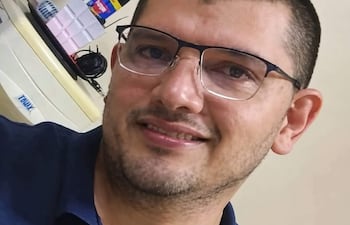 Hombre con gafas y barba ligera, sonriendo en una oficina, con camiseta oscura y fondo de objetos diversos.