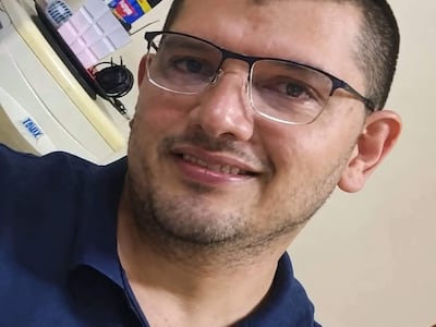 Hombre con gafas y barba ligera, sonriendo en una oficina, con camiseta oscura y fondo de objetos diversos.