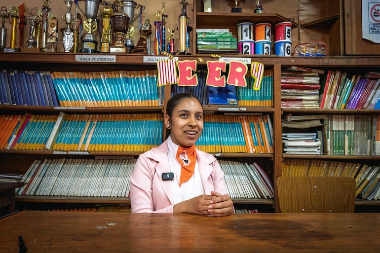 Thiara Pérez sonríe, viste blusa blanca y chaqueta rosa, en una biblioteca con estantes de libros y trofeos al fondo.