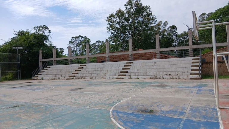 Cancha de baloncesto vacía con gradas de concreto y árboles al fondo bajo cielo nublado.
