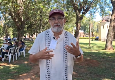 Hombre mayor con gorra burdeos y camisa clara habla enérgicamente, mientras personas escuchan sentadas en un entorno natural.
