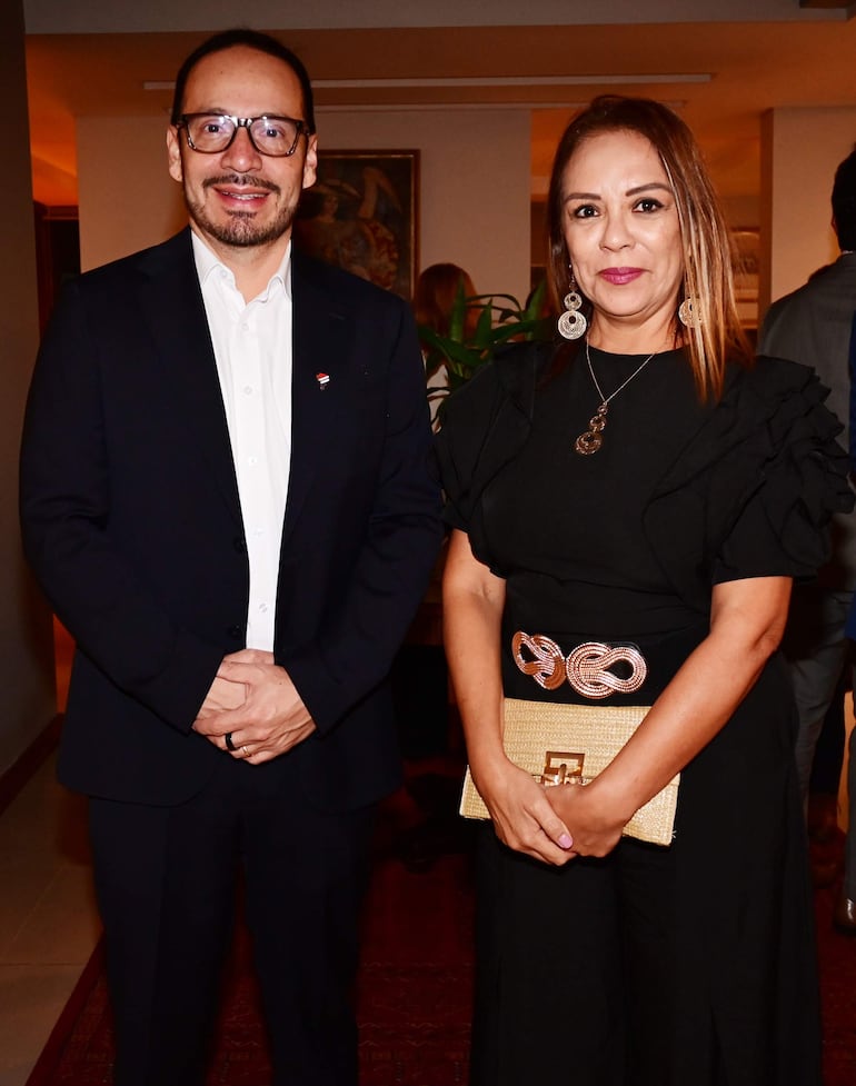 Hombre de traje oscuro y mujer en vestido negro sonríen en evento social decorado con tenue iluminación.