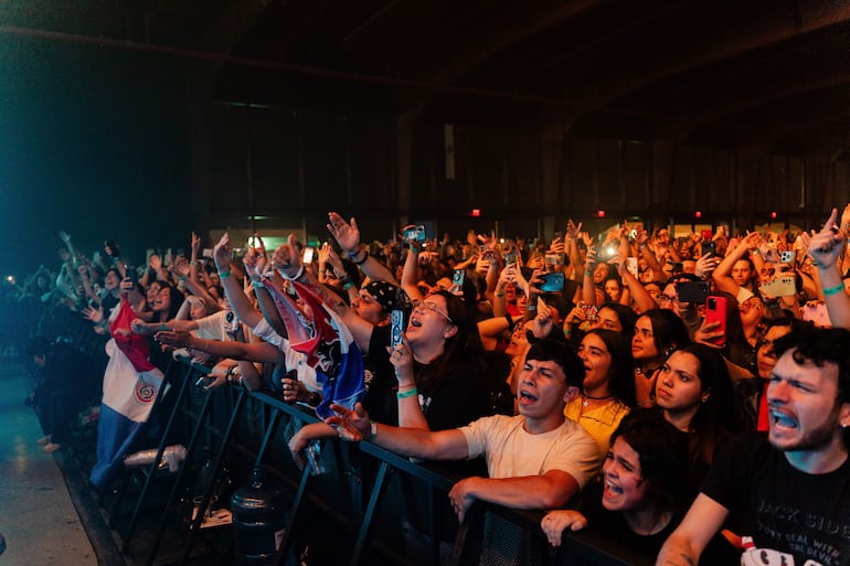 Multitud entusiasta en concierto, donde numerosos asistentes levantan las manos y sostienen teléfonos, rodeados de banderas paraguayas.