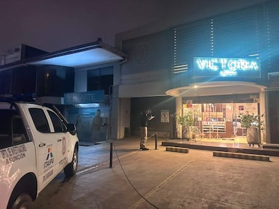 Agente policial de pie con cuaderno en mano frente a la joyería Victoria, con vehículo policial iluminado en un entorno urbano nocturno.