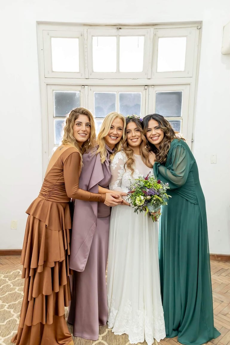 Menchi Barriocanal en vestido blanco con ramo de flores, sonriente, acompañada de tres mujeres elegantes en un entorno luminoso.