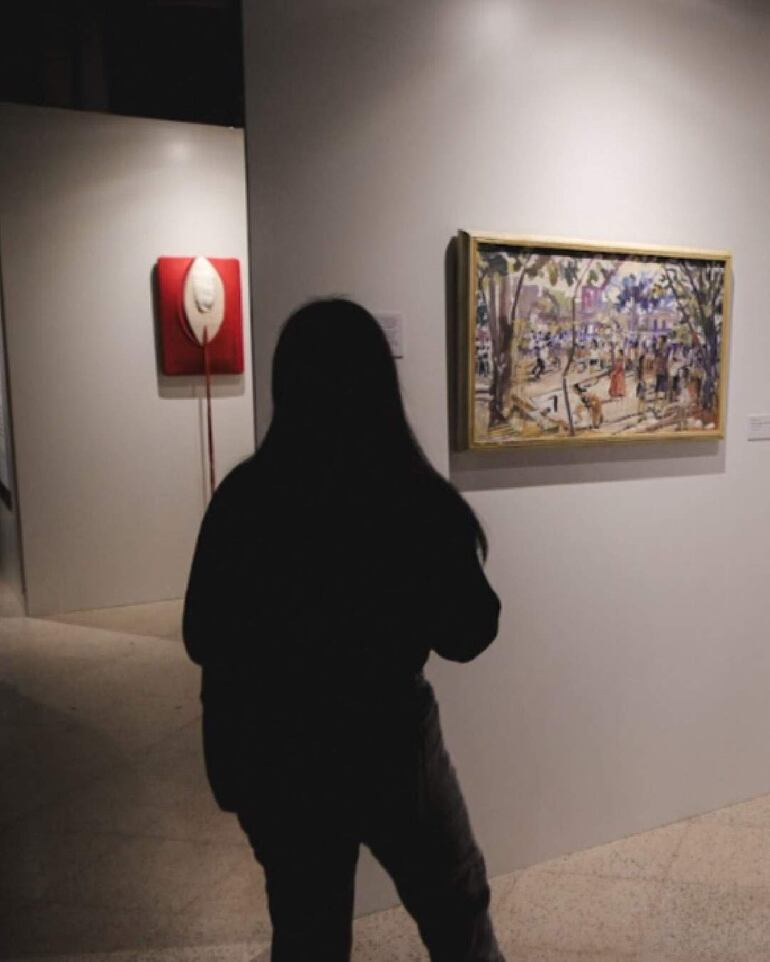Persona de espaldas en chaqueta oscura observa una pintura enmarcada en galería con paredes blancas y suelo claro.
