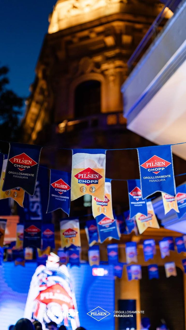 Banderines de Pilsen Chopp decoran un evento festivo al aire libre con iluminación nocturna y un fondo arquitectónico icónico.