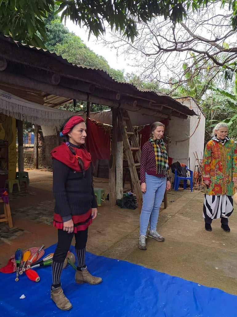 Tres mujeres: una con bufanda roja, otra en suéter oscuro y la última con pantalón a rayas, en un entorno rústico.