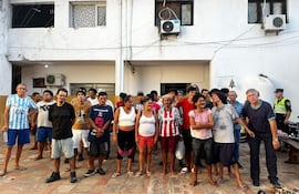 Grupo de personas en espacio urbano, algunas con camisetas de fútbol. Hombre con camiseta celeste observa mientras policía se encuentra al fondo.