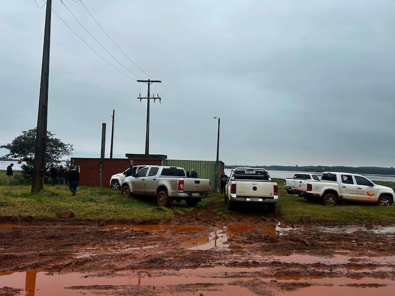 Grupo de personas conversando cerca de edificio de ladrillo, varias camionetas blancas estacionadas en terreno de barro y nublado.