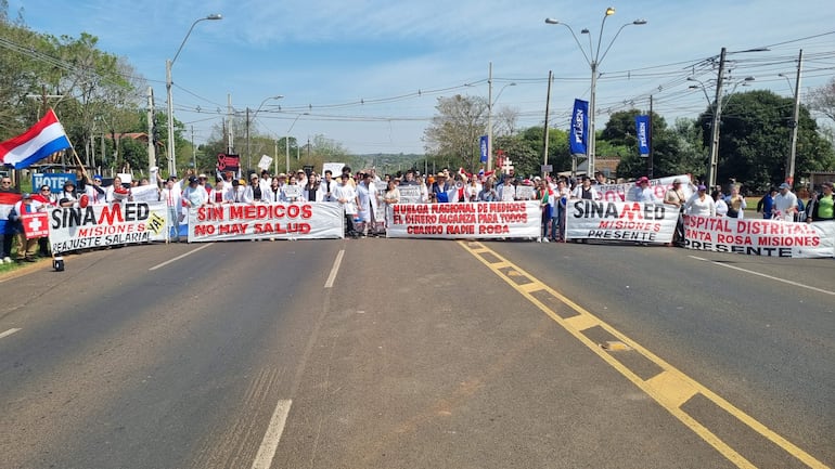 Cerca de 100 médicos en batas sostienen pancartas en plena protesta, destacándose la bandera paraguaya en el fondo.