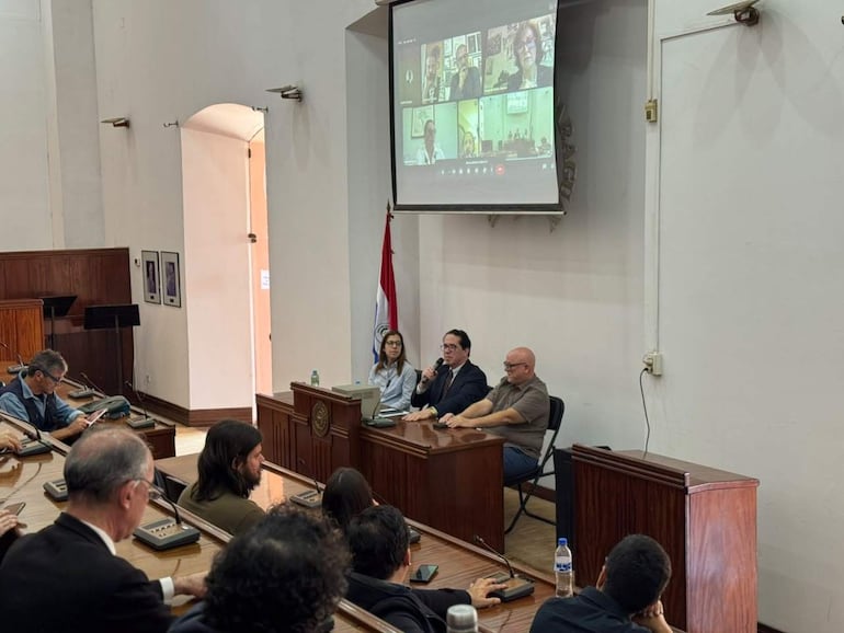 Mujer moderando, hombre con micrófono y otra persona observando en un auditorio con diseño sobrio y pantalla grande.