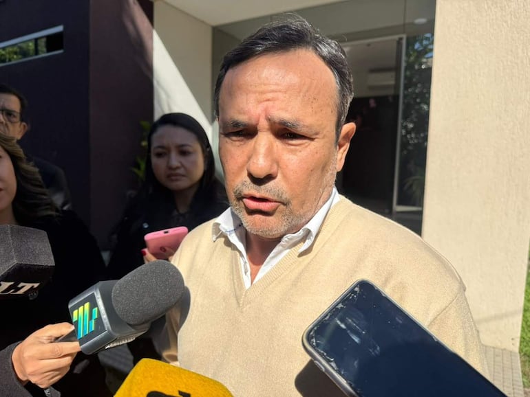 Emiliano Fernández en prenda beige, con expresión concentrada, responde a preguntas de periodistas frente a un edificio moderno.