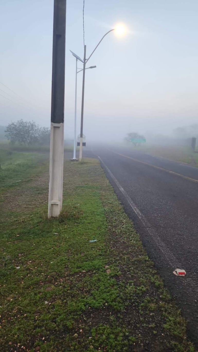 Carretera rural en niebla densa con un poste de luz encendido a la izquierda y carteles visibles en la bruma.