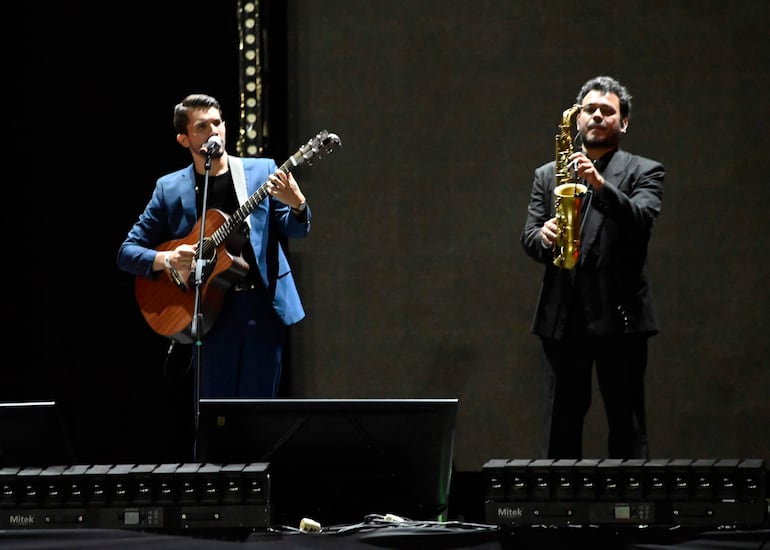 Montaner con traje azul y guitarra acústica canta al micrófono, acompañado por un saxofonista también vestido elegantemente.