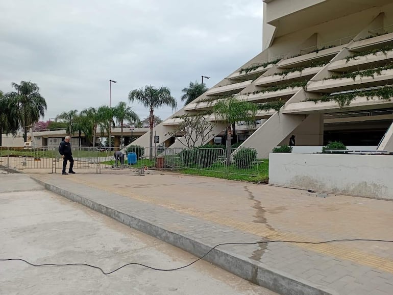 Dos trabajadores en el área del estacionamiento; uno supervisa de pie mientras el otro repara el suelo. Entorno con vegetación y arquitectura moderna.