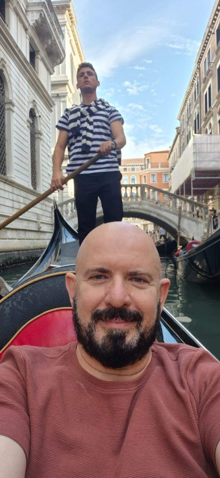 Hombre sonriente en primer plano con camiseta marrón, mientras otro rema en fondo, rodeados de canales venecianos.