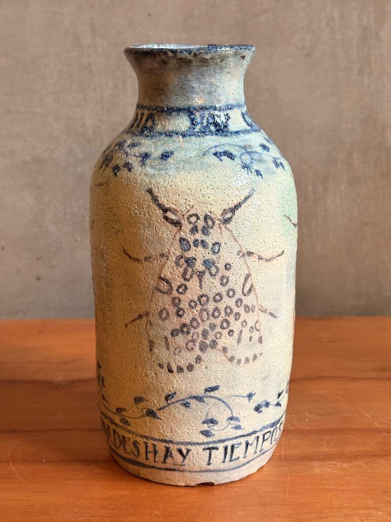 Vasija de cerámica beige con detalles azulados, exhibida sobre superficie de madera clara en un ambiente interior.