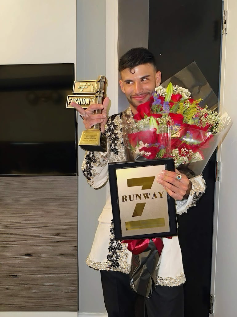 Simón Villalba sostiene un trofeo de 'Best Model' y un ramo de flores, luciendo satisfecho en un entorno neutro.