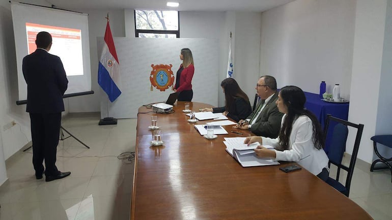Mujer de pie en blazer rojo explica a un grupo en salón de conferencias, rodeados de documentos y una bandera de Paraguay.