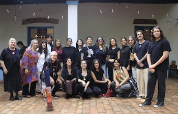Diecisiete artistas, principalmente mujeres, posan sonrientes en un ambiente acogedor con piso de ladrillo, vestidos en su mayoría de oscuro.