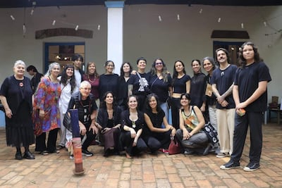 Diecisiete artistas, principalmente mujeres, posan sonrientes en un ambiente acogedor con piso de ladrillo, vestidos en su mayoría de oscuro.