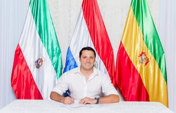 Hombre de camisa blanca escribiendo con bolígrafo, sentado frente a tres banderas: Paraguay, Itapúa y Coronel Bogado, con expresión amigable.