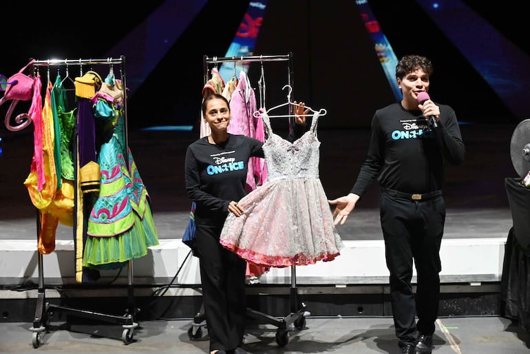 Hombre con micrófono y camiseta negra presenta vestuarios, mientras una mujer sostiene un vestido, ambos en un escenario colorido.