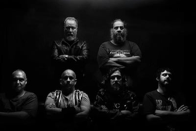 Seis hombres, tres con barba, en ambiente oscuro; algunos visten camisetas de bandas, mientras otros llevan chaquetas de cuero.